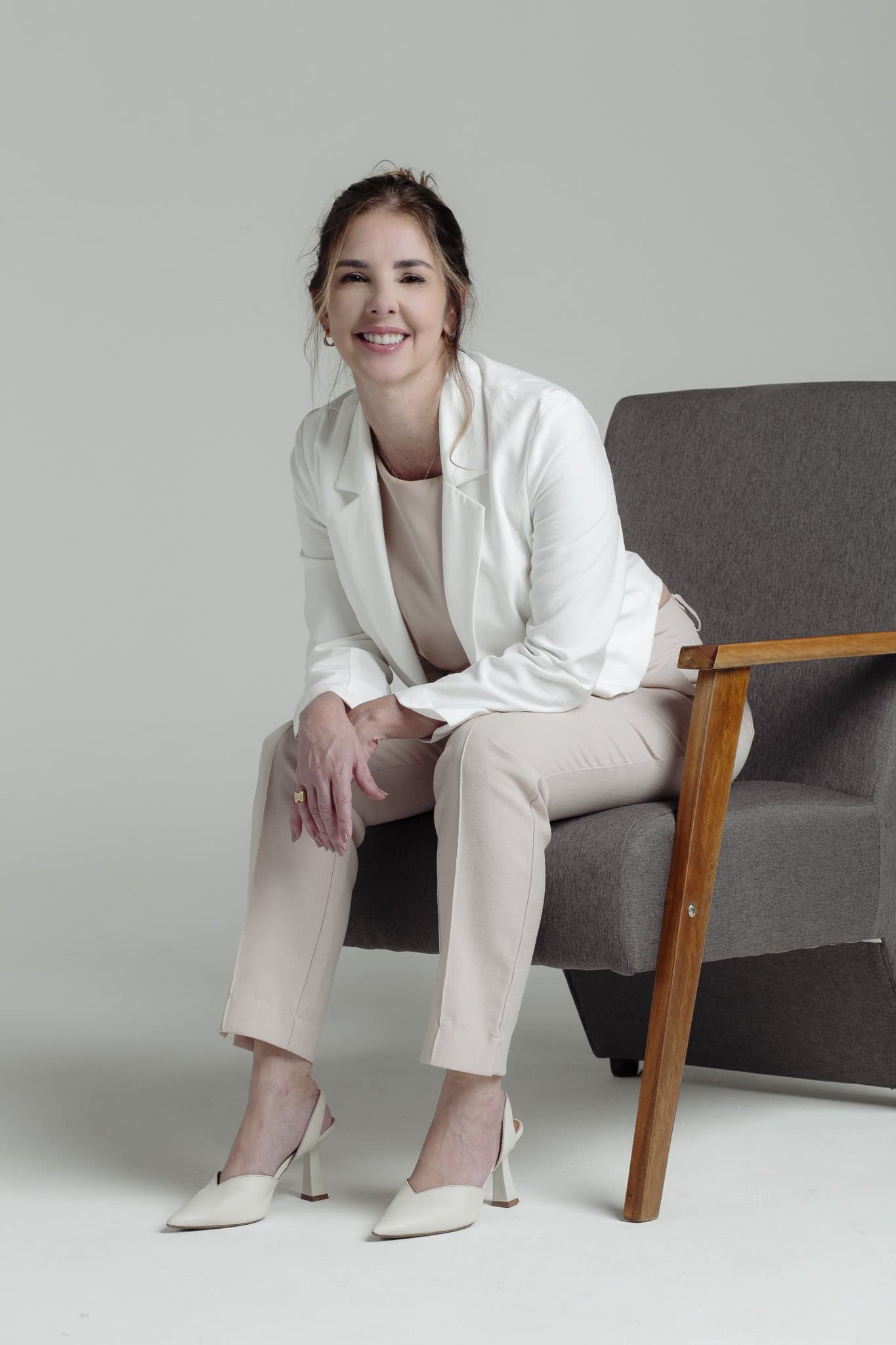 Anna Braga sentada em uma poltrona, de terno claro.